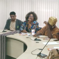 Um grupo de pessoas está sentado diante de um balcão branco curvo, incluindo duas mulheres (uma delas falando ao microfone) e um homem de terno. Um grupo de pessoas está sentado diante de um balcão branco curvo, incluindo duas mulheres (uma delas falando ao microfone) e um homem de terno.