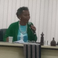 Uma mulher, usando blusa azul e blusa de frio verde, está sentada à mesa segurando um microfone. Uma mulher, usando blusa azul e blusa de frio verde, está sentada à mesa segurando um microfone.