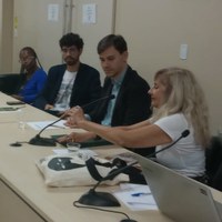 Um grupo de pessoas sentado à mesa com microfones, incluindo uma mulher de óculos e camisa azul ao fundo, um homem com camiseta branca estampada e um homem com camisa azul, ambos usando blazer, e uma mulher com camiseta branca ajusta um microfone. Em primeiro plano, há um notebook. Um grupo de pessoas sentado à mesa com microfones, incluindo uma mulher de óculos e camisa azul ao fundo, um homem com camiseta branca estampada e um homem com camisa azul, ambos usando blazer, e uma mulher com camiseta branca ajusta um microfone. Em primeiro plano, há um notebook.