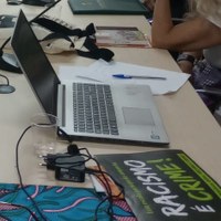 Um notebook sobre uma mesa, próximo a um copo de plástico com pouca água. É possível ver os braços de uma pessoa, um com relógio de pulseira preta, e um cartaz preto com destaque para o texto 'Racismo é crime!' em branco e verde claro, entre outras frases. Um notebook sobre uma mesa, próximo a um copo de plástico com pouca água. É possível ver os braços de uma pessoa, um com relógio de pulseira preta, e um cartaz preto com destaque para o texto 'Racismo é crime!' em branco e verde claro, entre outras frases.