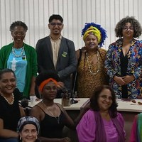Um grupo de pessoas posando para uma foto. A partir da esquerda superior, uma mulher com camisa azul e colar, um homem de terno, uma mulher com vestido amarelo e colar, uma mulher com jaqueta colorida, e, agachadas, estão uma mulher com camisa preta, uma mulher com regata preta, uma mulher com camisa rosa, além do rosto de uma mulher em primeiro plano. Um grupo de pessoas posando para uma foto. A partir da esquerda superior, uma mulher com camisa azul e colar, um homem de terno, uma mulher com vestido amarelo e colar, uma mulher com jaqueta colorida, e, agachadas, estão uma mulher com camisa preta, uma mulher com regata preta, uma mulher com camisa rosa, além do rosto de uma mulher em primeiro plano.