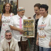 Pessoas em vernissage com artista Pedro Galvão segurando quadro ladeado por Selma di Medeiros e corpo discente de Jovens Aprendizes