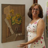 Artista plástica Selma di Medeiros sorrindo ao lado de tela com pintura de ipê amarelo e figuras humanas