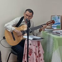 Músico e servidor Luis Libâneo sentado tocando violão e cantando em microfone durante evento cultural