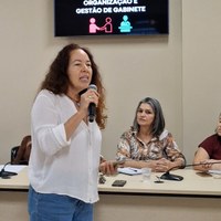 Servidora da Escola Legislativa utiliza microfone com tela de apresentação sobre gestão de gabinete ao fundo