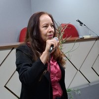 Servidora da Escola Legislativa, com cabelos longos castanhos blazer preto e blusa rosa segura microfone em oficina de oratória política na CMG