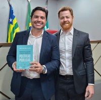 Procurador Lucas Velasco e um homem em pé segurando livro Racionalidade Legislativa e Produção Normativa das Políticas Públicas à frente de bandeiras oficiais