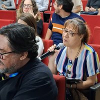 Público participante em auditório com mulher em primeiro plano falando ao microfone