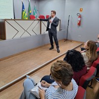 Procurador Lucas Velasco em pé ministrando palestra com auxílio de projeção para público em auditório