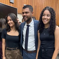 Vereador Romário Policarpo sorri ao lado de 2 estudantes em ambiente com painéis de madeira