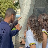 3 estudantes em visita guiada tocam superfície de monumento artístico na área externa da Câmara Municipal de Goiânia