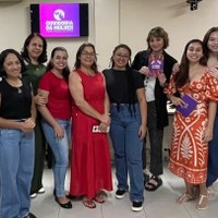 Mulheres em visita institucional posam à frente de monitor com logotipo da Ouvidoria da Mulher da Câmara Municipal de Goiânia